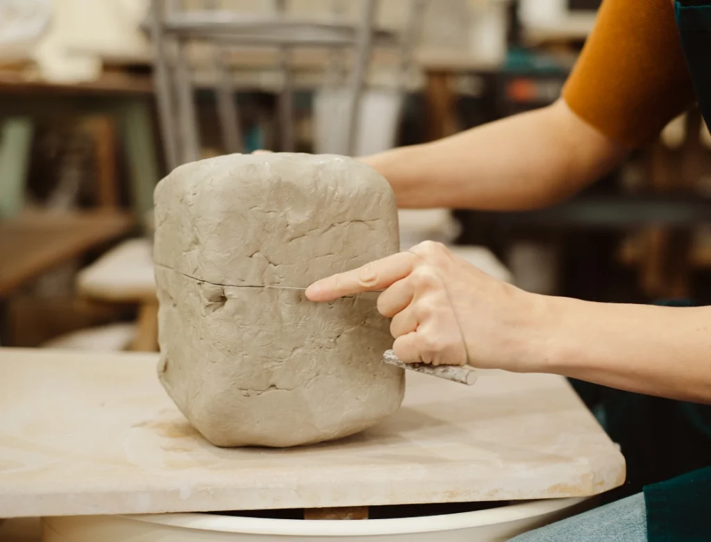 Person sculpting a rectangular clay block with hand tools in a pottery studio.