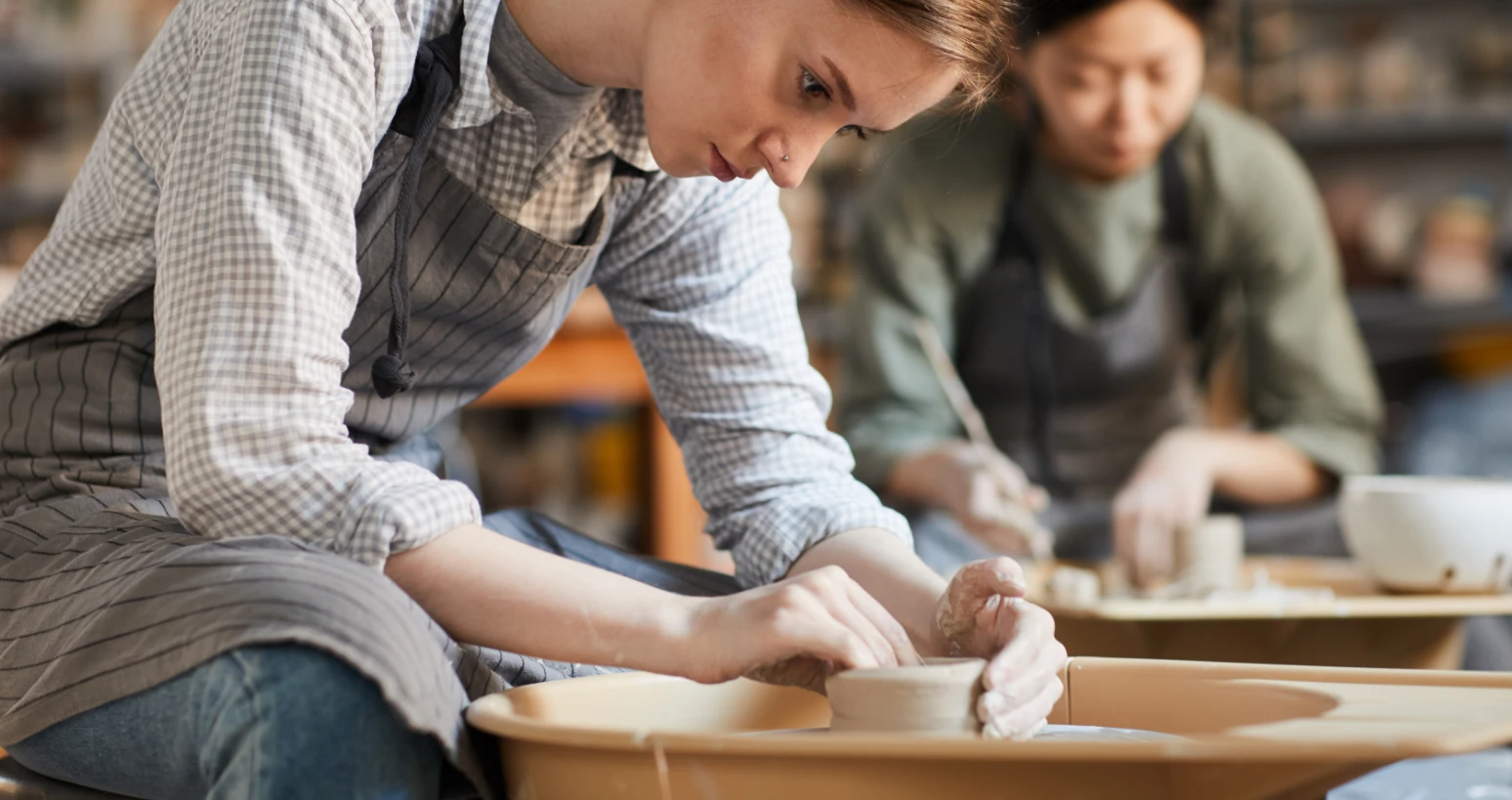 A woman shaping clay on a pottery wheel in a ceramics studio, with another person working in the background.