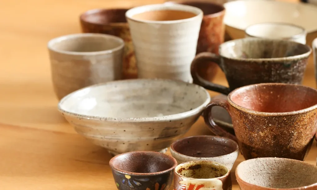 A collection of handmade ceramic cups and bowls in various earth tones arranged on a wooden surface.