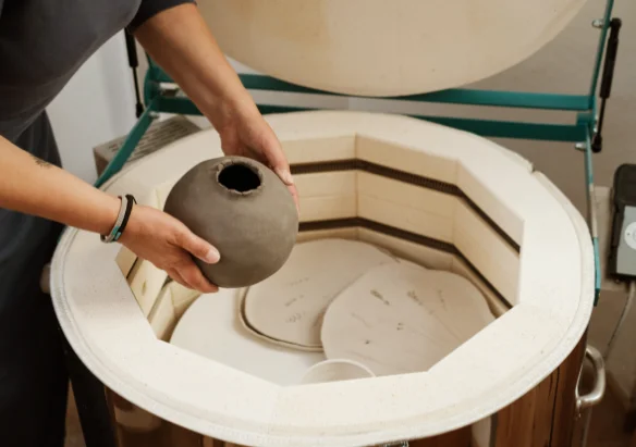 Person placing a round ceramic pot inside a pottery kiln for firing.
